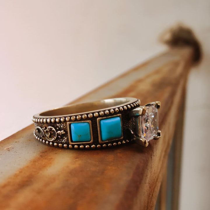 Square turquoise and zircon ring with decorative band on rustic surface.