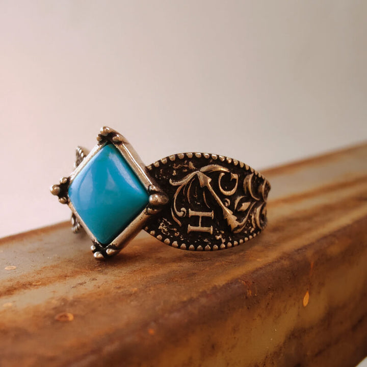 Floral engraved silver ring featuring a square turquoise stone on a wooden surface.