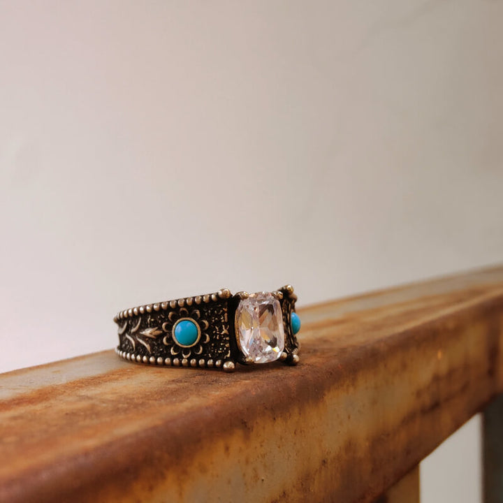 Vintage engagement ring with clear stone and turquoise accents on ornate metal band.