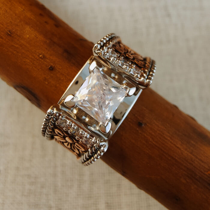 Two-tone floral wide band ring featuring a square zircon stone on wooden background.