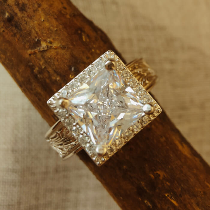 Square diamond ring with zircon gem and flower engravings, set on a rustic wooden surface.