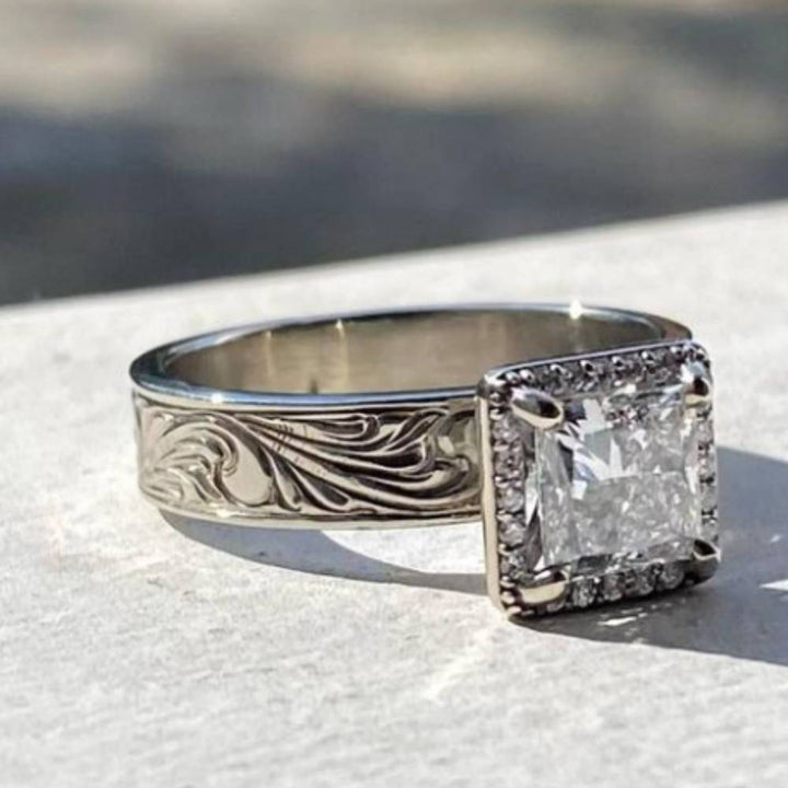 Square diamond ring with floral engravings and zircon gem on stone surface.